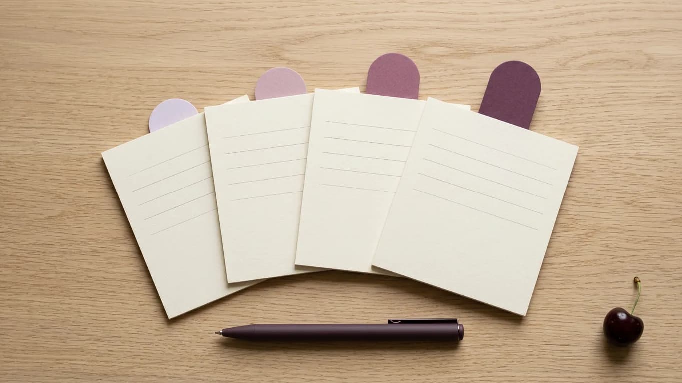 Four small sheets of pale paper fanned out on light oak, each with a few faint ruled lines and a single plum tab of a different height at its top edge, with a matte dark-plum pen resting below them