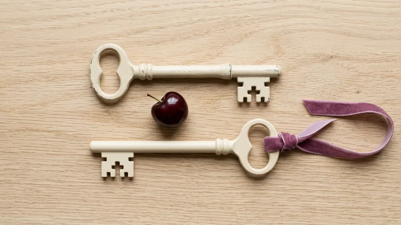Two nearly identical pale cream door keys lying side by side on light oak, one slightly worn and one new, with a single dark cherry between their heads and a soft plum ribbon threaded through one key's ring